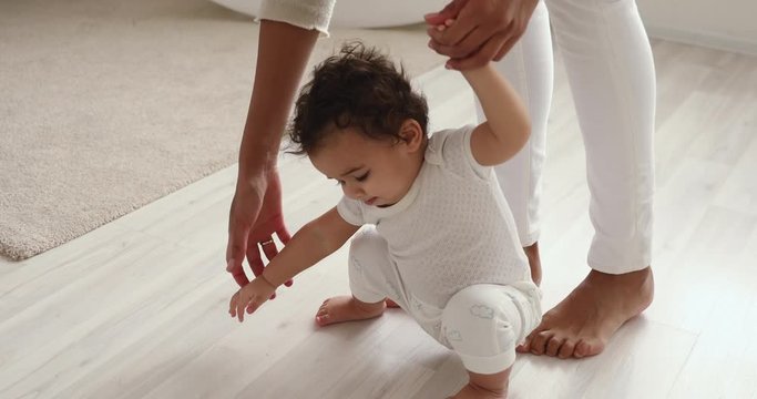 Cute barefoot small mixed race baby girl learning to walk holding mom hands. Funny adorable african infant toddler child daughter playing with mum making first step on warm heated floor. Close up view