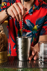 Bartender in a hawaiian shirt prepairing a cocktail in a mixing glass, a coconut and a rocks glass...