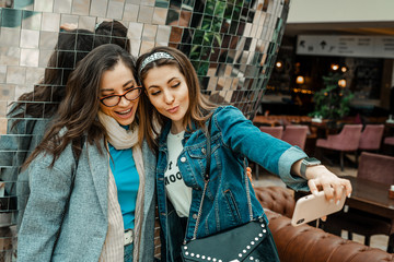 Fototapeta premium Trade, buyers. Two beautiful girls make purchases in a shopping center, go shopping. They look at the smartphone, take a selfie. The joy of consumption.