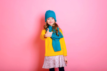 Blonde kid child girl is dressed in a warm sweater, scarf and beanie on a pink background. Fight the cold. Winter and autumn seasons. Concept of children's health, vitamins, medicines.