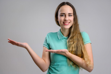 Fototapeta premium Photo of a pretty girl with long hair in a turquoise t-shirt on a white background. Model shows her hands to the sides, demonstrating the beauty of a smile