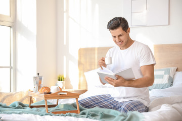 Morning of handsome young man reading book during breakfast in bed