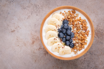 A bowl with with yogurt, granola, bananas and blueberry on beige concrete background with copy space