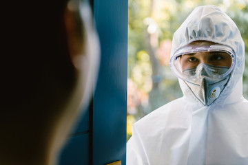 Health care worker in full virus protective suit standing in a front door of asian home. With copy space.