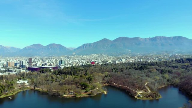 City buildings and great park with trees forest and calm lake, mountains and blue sky background in Tirana, Albania