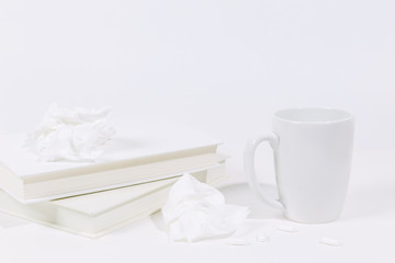 Cold season and flu concept flat lay with used tissues, ceramic mug, pills, and stacked pile of books. Monochromatic white color story.