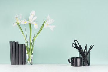 Light spring school workplace with black stationery, coffee cup, books, fresh white iris in vase on green mint menthe wall and white wood desk.