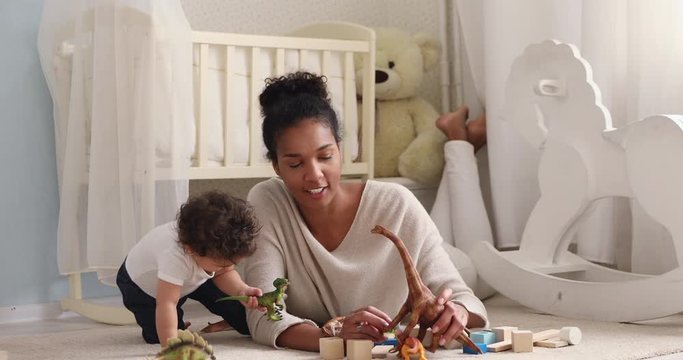 African family young adult mommy and cute small baby girl daughter playing toys relaxing on bedroom carpet. Happy mother or nanny teaching infant child boy son learning dinosaurs enjoy home activity.