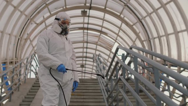 Epidemiologist in protective uniform sprays liquid chemicals on handrails in public places