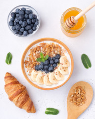 Breakfast consisting of yogurt, granola, bananas, blueberry and croissant on white concrete background with mint leaves