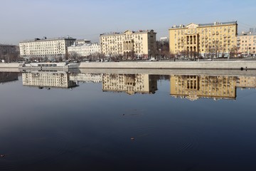 Obraz premium Buildings of the Frunze Embankment in Moscow are reflected in the smooth blue water of the Moscow River. Early spring, sunny day.