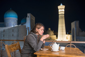 European she-tourist drink tea in front of Great Minaret of the Kalon, Kalon (Kallan) Mosque and Mir-i Arab (Miri Arab) Madrasah. Bukhara, Uzbekistan, Central Asia.