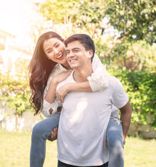 Portrait of cute romantic caucasian guy in checkered shirt, dreamy asian lady rides him on rear. Leisure, chill happiness, lawn stroll, relax, romance lifestyle, well dressed partners posing