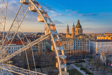 Naklejka premium Budapest, Hungary - Aerial view of the ferris wheel at Elisabeth Square (Erzsebet ter) at sunset with warm colors, St. Stephen's Basilica and blue sky at background