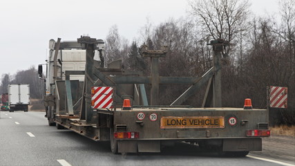 Long vehicle heavy oversized empty semi truck with low-frame semi-trailer trawl drive on suburban asphalted road at spring day, close up rear-side view – Logistics, oversize transportation trucking