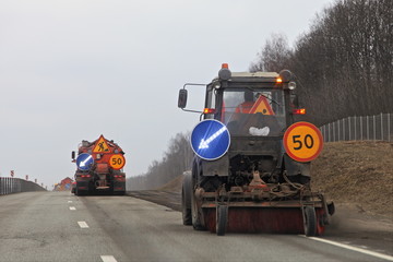 Road cleaning in Russia, asphalt sweeper tractor cleans spring roadside on trees background,...