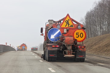 Naklejka premium Russian orange asphalt sweeper truck on spring road on trees background, municipal highway cleaning service