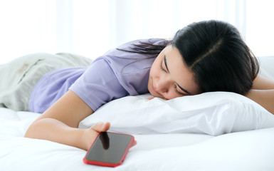 Beautiful Asia woman lying on the bed and holding smartphone in her hand. sleep in the white bedroom, focused on women. Feeling tried. Social addicted, modern life style and relaxation concept.