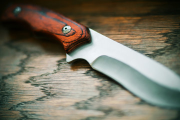 A sharp steel hunting knife with a wooden handle lies on a wooden table, illuminated by the light.