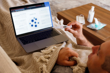 Close up shot of adult man working at home due to coronavirus quarantine concept. Male sitting on couch with laptop, measuring body temperature. Antiseptic gel and wet wipes on background, copy space.