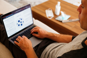 Close up shot of adult man working at home due to coronavirus quarantine concept. Male sitting on couch with laptop, measuring body temperature. Antiseptic gel and wet wipes on background, copy space.