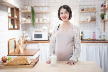 Portrait beautiful young asian pregnant woman drink milk glass standing in kitchen at home.Health care and love of pregnancy mother’s day concept.