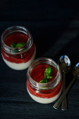 Tasty fragrant panacotta with strawberry compote decorated with fresh basil in glass jars next to silver spoons on a black background. View from above.