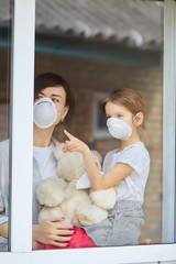 Mother with daughter child in mask looking from window, coronavirus.