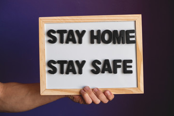 inscription stay home stay safe on a white chalk board in a man;s hand opposite violet wall