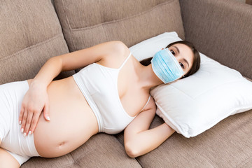 Obraz na płótnie Canvas Pregnant woman is wearing protective medical mask to protect herself and future baby from coronavirus. Mother is lying on the couch at home. Global quarantine concept