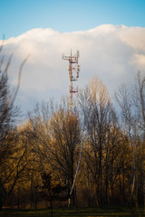 A radio tower with some huge clouds in the background and trees in the foreground.