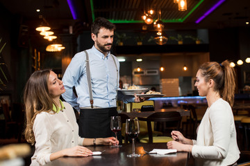 Smiling young female friends at a restaurant with waiter serving dinner