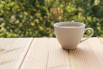 white cup with coffee on a light wooden table against a green summer garden, close-up, copy space