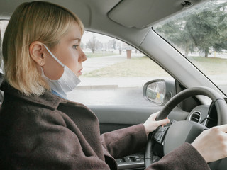 Young woman driving a car while wearing a white protective face mask as precaution against the covid-19 virus pandemic. Coronavirus lifestyle adaptation.