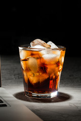 glass of cola with ice and laptop on grey stone table on black background