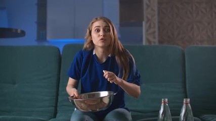 Portrait shot of the young good looking woman sitting in front of TV on the sofa with popcorn and beer while watching sport game and his team loosing. Indoors.