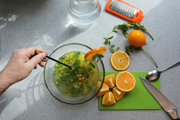 The process of making homemade lemonade. Lemon, orange, mint are on the gray table. A man's hand mixes ingredients in a transparent bowl. Concept of healthy food, lifestyle and diet. Selective focus.