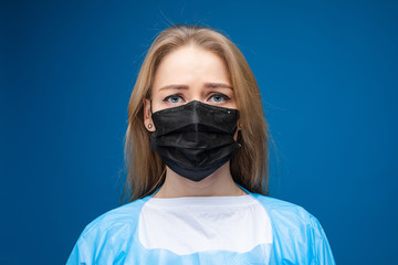 Young beautiful caucasian female in blue medical gown and with black medical mask on her face looks on the camera and is sad