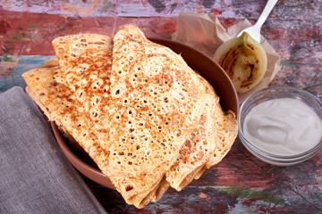 pancakes on yeast lie on a dish, next to a fork, sour cream, textile napkin