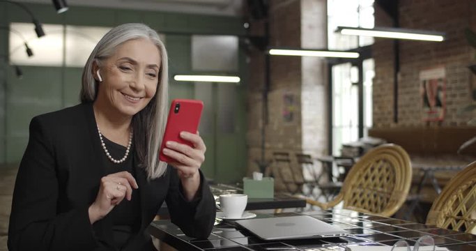 Front view of elegant lady businessowner in earpods having videocall on smartphone. Modern middle aged woman sitting in cafe and communicating using videoconference. Concept of tech.