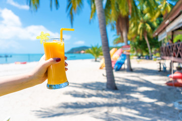 First-person view. Girl holds a glass cup of cold mango fresh on the background of a sandy tropical beach. White sand and palm trees. Fairytale vacation