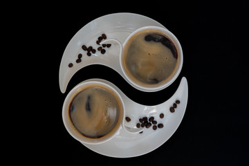 Two white cups with black coffee are standing on two white plates with coffee beans against black background, top view