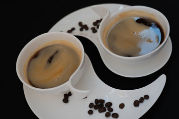 Two white cups with black coffee are standing on two white plates with coffee beans against black background, closeup side view