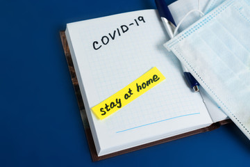 text coronavirus, stay at home, office there written on a Notepad by hand. the view from the top. mask and ballpoint pen with Notepad on a blue background
