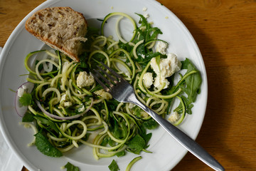 Unfinished salad on a wooden table. A cold appetizer of zucchini noodles, with fresh herbs and feta.On a white plate lies a piece of bread and a fork.Vegetarian version of the dish. Healthy nutrition.