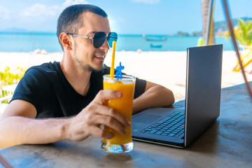 The guy works at a laptop, sitting in a beach bar and drinks a mango shake. Successful business. Remotely work.