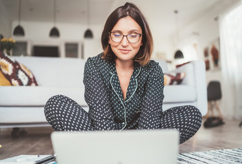Smiling  Businesswoman dressed pajamas typing notebook keyboard chatting with colleagues sitting cross-legged on living room floor office with laptop.Distance work in worldwide quarantine time concept