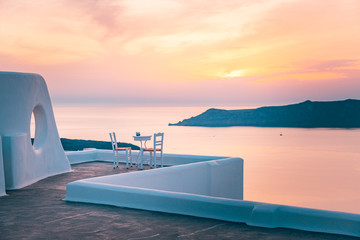White architecture on Santorini island, Greece. Outdoor restaurant under a fantastic sunset landscape, chairs for couple. Romantic sunset view, orange sky and clouds. Summer vacation, honeymoon travel