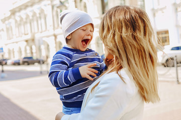 A small child laughs merrily in front of a young mother in her arms while walking around the city center. A clear sunny day.