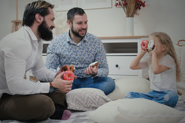 Little cute girl with her two dads sitting on the floor in apartment and play with glazed donuts. Homosexual couple with their daughter make funny faces with donuts